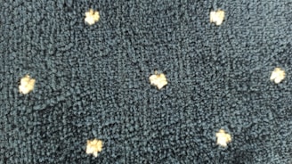 A textured dark fabric with a pattern of small, light-colored, star-like shapes evenly distributed across the surface. The texture appears plush and thick, indicating a potential use in upholstery or carpeting.