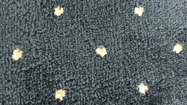 A textured dark fabric with a pattern of small, light-colored, star-like shapes evenly distributed across the surface. The texture appears plush and thick, indicating a potential use in upholstery or carpeting.