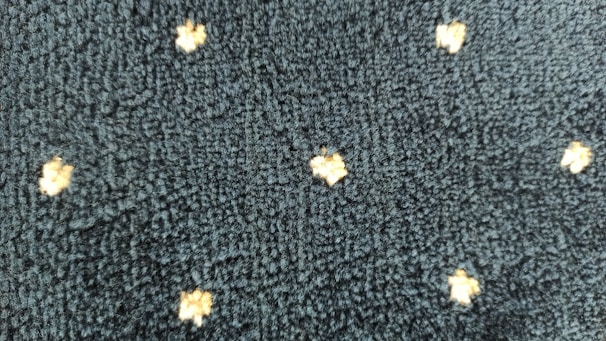 A textured dark fabric with a pattern of small, light-colored, star-like shapes evenly distributed across the surface. The texture appears plush and thick, indicating a potential use in upholstery or carpeting.