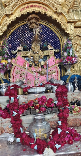 An ornate Hindu altar featuring a deity adorned with vibrant pink garments and elaborate gold jewelry. The background is deep blue with gold embellishments, and peacock motifs are present. The altar is surrounded by colorful floral arrangements, with a prominent red garland. Silver vessels and offering items are laid out in front.