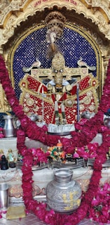 An intricately decorated idol adorned with a lavish red garment, embedded with golden and silver embellishments. The background features a deep blue backdrop with sparkling elements. The scene is framed by an ornate golden arch, and a garland of vibrant red flowers drapes symmetrically. Additional objects include silver vessels and small figurines arranged around the central idol.