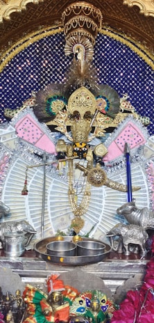 An ornate and intricately decorated shrine featuring a gold-toned deity figure adorned with peacock feathers and various colorful fabrics. It is surrounded by silver and metallic elements, including two animal figures on either side and ritual items such as bowls and a bell. The background is a deep blue with an array of small decorative lights.