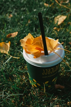 A paper cup with a black straw and a lid is placed on grass. The cup is decorated with a pattern of yellow leaves and writing. On the lid, a few large, bright yellow autumn leaves are resting.