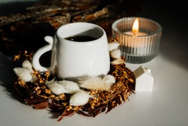 A white ceramic mug filled with a dark beverage is surrounded by dried floral arrangements and sits on a white surface. A lit candle in a ribbed glass holder emits a warm glow beside the mug. There is also a small, white decorative object resembling a house.