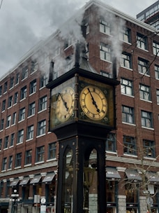 A steam-powered clock stands in front of a tall brick building. Smoke is visibly billowing from the top of the clock, which features two round faces with Roman numerals. The building has multiple windows and awnings, contributing to an urban cityscape.