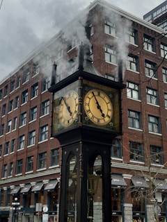 A steam-powered clock stands in front of a tall brick building. Smoke is visibly billowing from the top of the clock, which features two round faces with Roman numerals. The building has multiple windows and awnings, contributing to an urban cityscape.