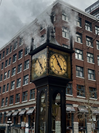 A steam-powered clock stands in front of a tall brick building. Smoke is visibly billowing from the top of the clock, which features two round faces with Roman numerals. The building has multiple windows and awnings, contributing to an urban cityscape.