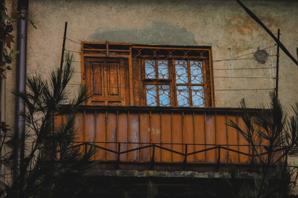 The image shows a balcony with a wooden railing and a window, both painted in a light brown color that contrasts with the dark gray concrete wall behind it. The window is adorned with a metal grate on its lower half, adding an interesting architectural detail to the scene. On either side of the balcony, there are two potted plants - one on each side.