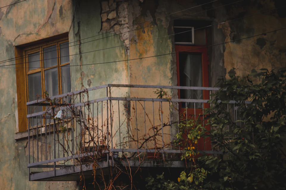 The image depicts an old, dilapidated building with a faded green facade and peeling paint. The building features a balcony on its second floor that is currently empty. A red door stands out against the muted colors of the structure. The perspective of the photo suggests it was taken from below, looking up at the building's facade.