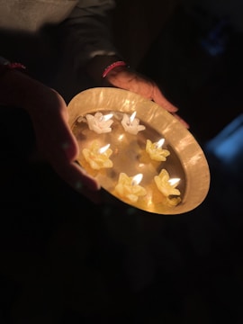 A pair of hands gently holds a golden tray containing several small, decorative candles shaped like flowers. The warm glow from the candles casts a soft light against the dark background, creating a serene and intimate atmosphere.