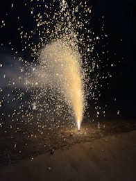 A fountain firework emits bright, sparkling light and smoke against a dark background. The golden sparks spread outwards and upwards, illuminating the surrounding area and creating a contrast with the night sky.