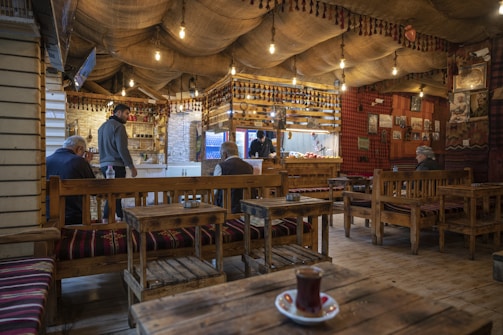 A cozy, traditional-style café with wooden benches and tables, decorated with ethnic patterns. Hanging light bulbs create a warm ambiance. A few people are seated and chatting, while a person stands near the counter. The walls are adorned with cultural decor and photographs.