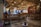 A cozy, traditional-style café with wooden benches and tables, decorated with ethnic patterns. Hanging light bulbs create a warm ambiance. A few people are seated and chatting, while a person stands near the counter. The walls are adorned with cultural decor and photographs.