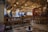 A cozy, traditional-style café with wooden benches and tables, decorated with ethnic patterns. Hanging light bulbs create a warm ambiance. A few people are seated and chatting, while a person stands near the counter. The walls are adorned with cultural decor and photographs.