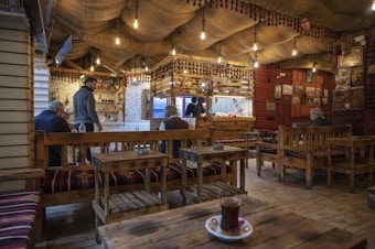 A cozy, traditional-style caf&eacute; with wooden benches and tables, decorated with ethnic patterns. Hanging light bulbs create a warm ambiance. A few people are seated and chatting, while a person stands near the counter. The walls are adorned with cultural decor and photographs.