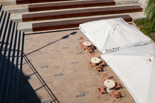 An outdoor area features several round tables with chairs arranged beneath large white umbrellas. A series of concrete steps lined with wooden beams leads to the terrace, which is paved with light stone tiles. The shadow of a lamp post stretches across the paving.