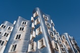 An architectural structure with a modern, innovative design featuring a wavy facade made of reflective metallic panels. Numerous windows with open shutters are distributed unevenly across the building's surface. The structure is set against a clear blue sky.