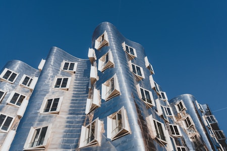 An architectural structure with a modern, innovative design featuring a wavy facade made of reflective metallic panels. Numerous windows with open shutters are distributed unevenly across the building's surface. The structure is set against a clear blue sky.