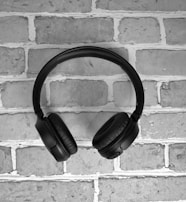 A pair of black over-ear headphones is positioned against a light-colored brick wall, highlighting the headphones' plush ear cushions and sleek design.