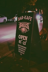 A sidewalk signboard placed at night in an urban setting, promoting a bar-cafe. The text is in a clear white color with details such as opening hours and types of drinks offered. Cars and trees are visible in the dimly lit background, creating a cozy, urban nighttime atmosphere.
