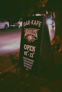 A sidewalk signboard placed at night in an urban setting, promoting a bar-cafe. The text is in a clear white color with details such as opening hours and types of drinks offered. Cars and trees are visible in the dimly lit background, creating a cozy, urban nighttime atmosphere.