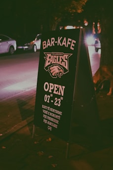 A sidewalk signboard placed at night in an urban setting, promoting a bar-cafe. The text is in a clear white color with details such as opening hours and types of drinks offered. Cars and trees are visible in the dimly lit background, creating a cozy, urban nighttime atmosphere.