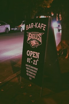 A sidewalk signboard placed at night in an urban setting, promoting a bar-cafe. The text is in a clear white color with details such as opening hours and types of drinks offered. Cars and trees are visible in the dimly lit background, creating a cozy, urban nighttime atmosphere.