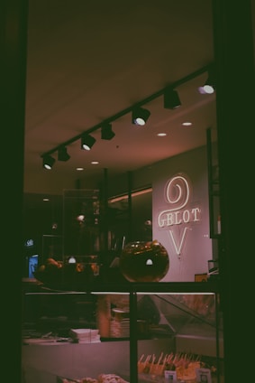 The image depicts the interior of a gelato shop with a neon sign displaying the word 'Gelato'. Various gelato flavors are visible in the display case with waffle cones and sticks. Overhead track lighting illuminates the area, contributing to a cozy ambiance.