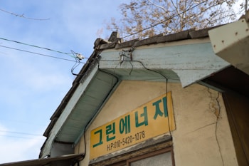 A weathered building facade with a yellow sign featuring text in Korean. The structure has a sloped roof with exposed wooden beams painted light blue and visible electrical wires. The background shows clear blue skies and some branches of trees without leaves.