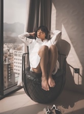 A person is sitting on a uniquely designed black chair, next to a large window. The individual is wearing a white shirt and has a relaxed posture, with legs drawn up and eyes closed. Sunlight is streaming in through the window, creating a warm and calming atmosphere. City buildings and mountains are visible outside the window.