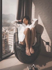 A person is sitting on a uniquely designed black chair, next to a large window. The individual is wearing a white shirt and has a relaxed posture, with legs drawn up and eyes closed. Sunlight is streaming in through the window, creating a warm and calming atmosphere. City buildings and mountains are visible outside the window.
