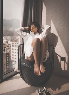 A person is sitting on a uniquely designed black chair, next to a large window. The individual is wearing a white shirt and has a relaxed posture, with legs drawn up and eyes closed. Sunlight is streaming in through the window, creating a warm and calming atmosphere. City buildings and mountains are visible outside the window.