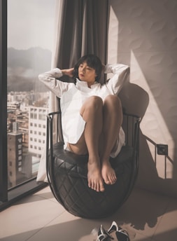 A person is sitting on a uniquely designed black chair, next to a large window. The individual is wearing a white shirt and has a relaxed posture, with legs drawn up and eyes closed. Sunlight is streaming in through the window, creating a warm and calming atmosphere. City buildings and mountains are visible outside the window.