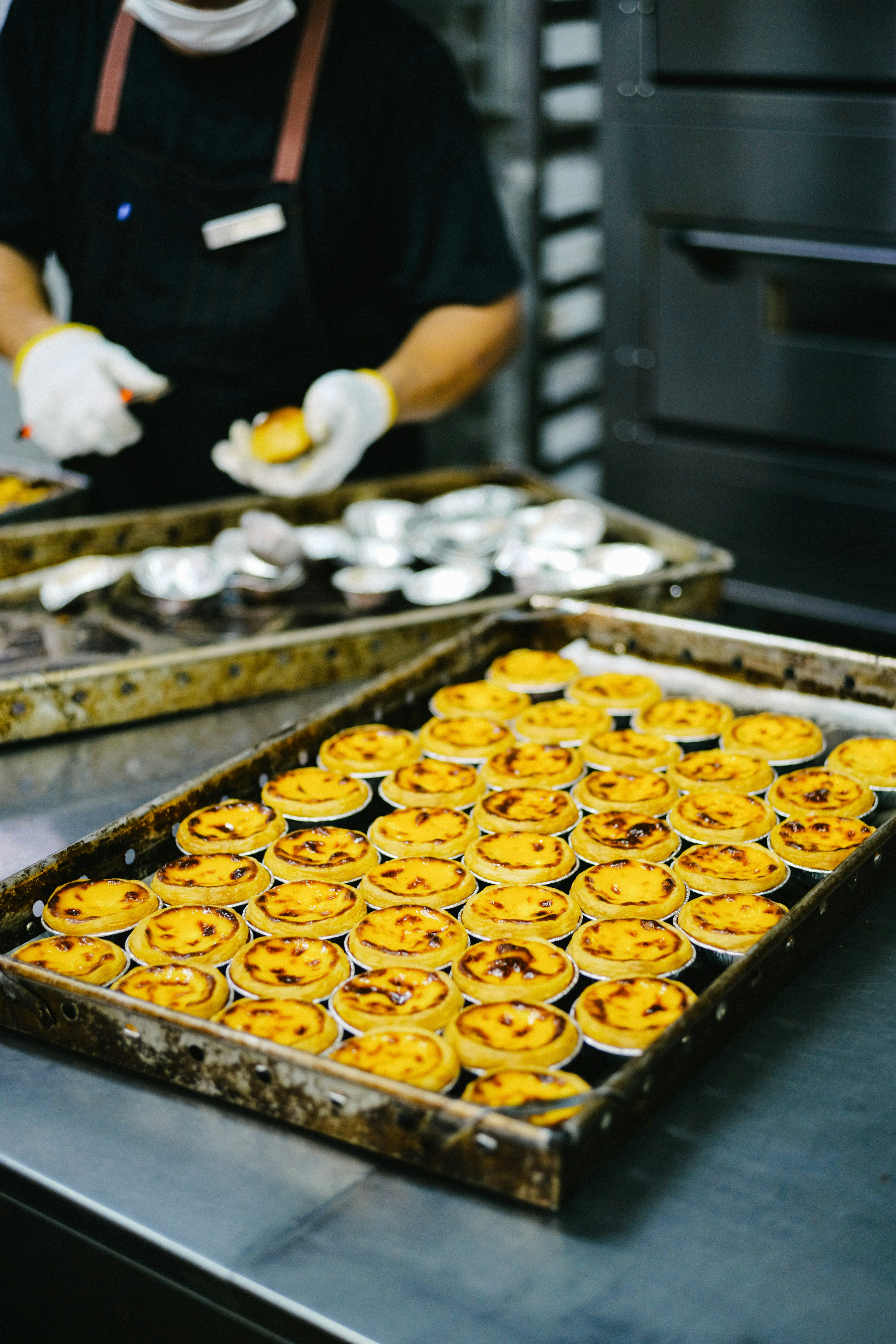 A baker in gloves arranges golden egg tarts on metal trays.