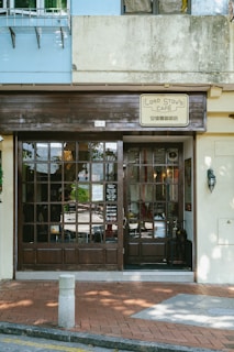 A quaint café facade with wooden framed glass doors and a sign reading 'Lord Stow's Café'. The exterior features a cream-colored wall and brick pavement, with some shadows cast by nearby trees.