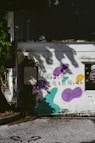 A weathered building exterior is adorned with graffiti art, including purple, teal, and yellow abstract shapes. A poetic message written in black text reads, "Sadness in my heart, I feel the best thing I could do." Shadows of tree branches and leaves decorate the wall, creating a serene yet contemplative atmosphere.