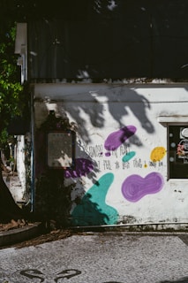 A weathered building exterior is adorned with graffiti art, including purple, teal, and yellow abstract shapes. A poetic message written in black text reads, "Sadness in my heart, I feel the best thing I could do." Shadows of tree branches and leaves decorate the wall, creating a serene yet contemplative atmosphere.