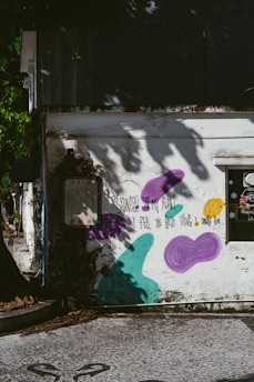 A weathered building exterior is adorned with graffiti art, including purple, teal, and yellow abstract shapes. A poetic message written in black text reads, "Sadness in my heart, I feel the best thing I could do." Shadows of tree branches and leaves decorate the wall, creating a serene yet contemplative atmosphere.