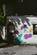A weathered building exterior is adorned with graffiti art, including purple, teal, and yellow abstract shapes. A poetic message written in black text reads, "Sadness in my heart, I feel the best thing I could do." Shadows of tree branches and leaves decorate the wall, creating a serene yet contemplative atmosphere.