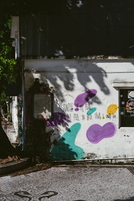 A weathered building exterior is adorned with graffiti art, including purple, teal, and yellow abstract shapes. A poetic message written in black text reads, "Sadness in my heart, I feel the best thing I could do." Shadows of tree branches and leaves decorate the wall, creating a serene yet contemplative atmosphere.