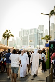 A busy urban street scene with people walking in a marketplace setting. Some individuals are wearing traditional Middle Eastern attire, while others are in casual clothing. Stalls with merchandise, a parking sign, and a backdrop of modern skyscrapers and palm trees contribute to a bustling city atmosphere.