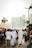 A busy urban street scene with people walking in a marketplace setting. Some individuals are wearing traditional Middle Eastern attire, while others are in casual clothing. Stalls with merchandise, a parking sign, and a backdrop of modern skyscrapers and palm trees contribute to a bustling city atmosphere.