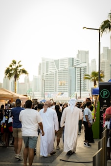 A busy urban street scene with people walking in a marketplace setting. Some individuals are wearing traditional Middle Eastern attire, while others are in casual clothing. Stalls with merchandise, a parking sign, and a backdrop of modern skyscrapers and palm trees contribute to a bustling city atmosphere.