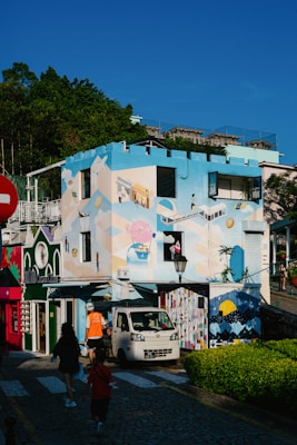 A vibrant, colorful building with artistic murals featuring geometric patterns and whimsical designs stands prominently under a clear blue sky. A small white truck is parked on the cobblestone street in front. Several people walk along the street, bordered by lush greenery. The building is flanked by other colorful small structures, adding to the lively atmosphere.