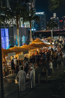 Nighttime street market bustling with activity. Stalls with yellow canopies line the sidewalk, illuminated by string lights wrapped around palm trees. Shoppers and visitors can be seen browsing the stalls, which are selling various goods. The backdrop includes a modern cityscape with high-rise buildings and bright neon signs.