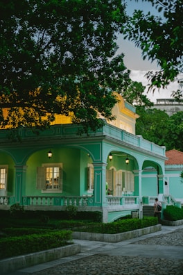 A quaint, colonial-style house painted in turquoise with white trim, surrounded by lush greenery. The house features a wrap-around porch with white balustrades and several lanterns hanging. Windows with shutters are open, revealing a warmly lit interior. A stone pathway and neatly trimmed bushes lead up to the entrance.