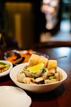 A bowl filled with clams, fresh herbs, a lemon wedge, and pieces of crusty bread is placed on a wooden table. The background features another dish and a warm, softly lit atmosphere, suggesting a cozy dining environment.