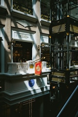 An indoor scene of a mall or shopping center featuring a luxury Ralph Lauren storefront with a Polo brand sign. The architectural design is ornate, with classical elements such as balconies, decorative light fixtures, and intricate moldings. Gold and black decorative motifs adorn the structure in the right side of the image.