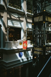 An indoor scene of a mall or shopping center featuring a luxury Ralph Lauren storefront with a Polo brand sign. The architectural design is ornate, with classical elements such as balconies, decorative light fixtures, and intricate moldings. Gold and black decorative motifs adorn the structure in the right side of the image.