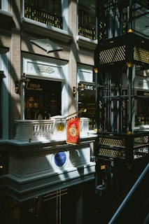 An indoor scene of a mall or shopping center featuring a luxury Ralph Lauren storefront with a Polo brand sign. The architectural design is ornate, with classical elements such as balconies, decorative light fixtures, and intricate moldings. Gold and black decorative motifs adorn the structure in the right side of the image.